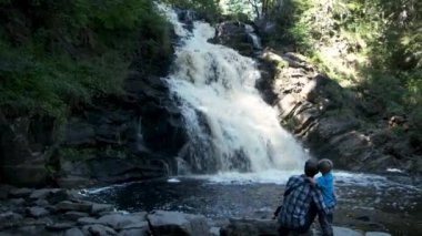 Baba ve oğul yürüyüşçüler dağ şelalesinin yanında oturuyorlar. Aktif aile açık havada, çocukla seyahat ve macera, bilinçli yaşam.