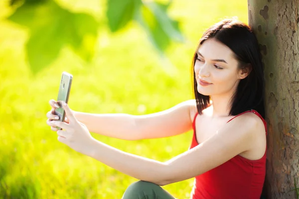 黑褐色头发的女孩用智能手机在树下自慰 — 图库照片