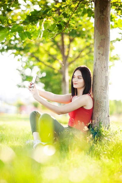 girl with dark brown hair using smartphone taking selfy under a tree