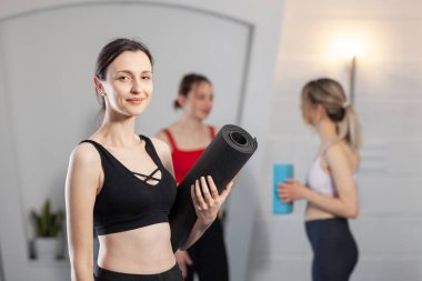 Yoga eğitmeni yoga minderiyle poz veriyor. Arka planda sohbet eden iki kız..