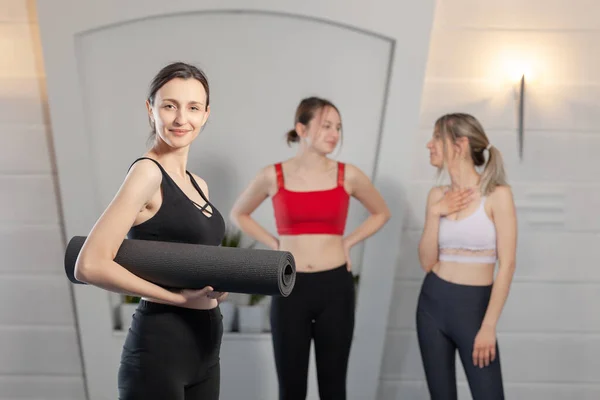 Yoga instructor posing with yoga mat. Two girls chatting in the background.