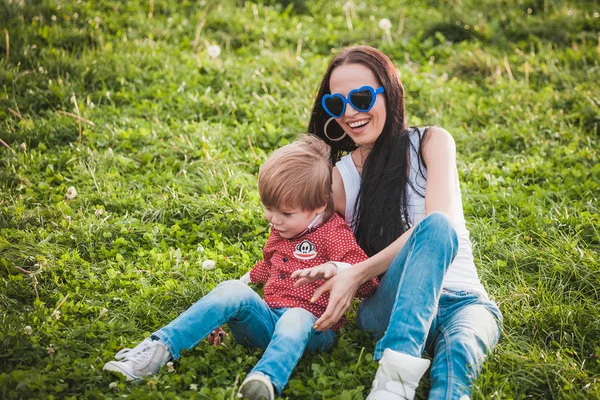 Lykkelig mor med sin lille søn sidder på græs i sommerdag - Stock-foto