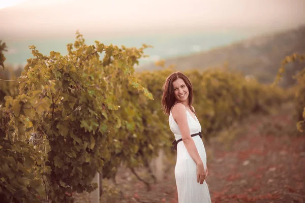 Улыбающаяся женщина в белом платье стоит в винограднике — стоковое фото