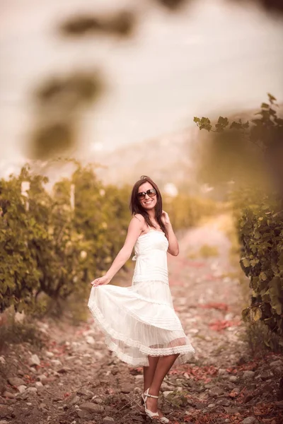 Mujer sonriente en vestido blanco de pie en el viñedo — Foto de Stock