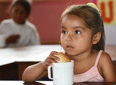 Iquitos, Peru - 12 Ekim 2015: Santa Ana köyü anaokulu yemek vakti. Genç bir kız bir rulo ve süt vardır.