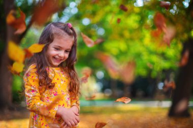 Küçük gülümseyen kız güneş parkında sonbahar yapraklarıyla oynuyor.