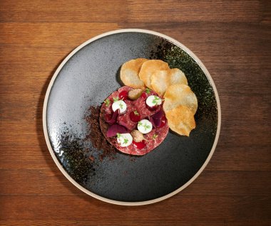 Üstünde patates kızartması ve sos olan kırmızı tartar eti siyah tabakta.