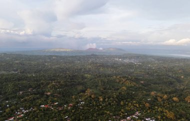 Kuş bakışı yemyeşil tepeleri ve bulutlu gökyüzünün altındaki uzak volkanik tepeleri gösterir. Doğanın günbatımındaki güzelliğini gösterir..