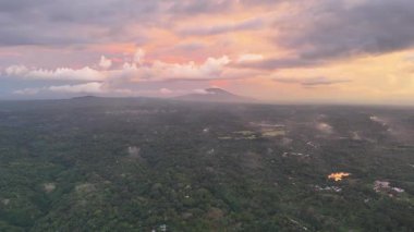 Canlı günbatımının, yeşil tepeleri aydınlatan ve sakin doğal ortamdaki uzak volkanın güzelliğinin tadını çıkarın..
