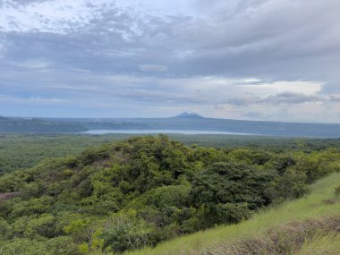 Lush greenery surrounds a serene lake with a volcano visible in the background under a cloudy sky during the day.