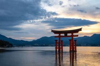 Itsukushima jinja Tapınağı 'nın ünlü yüzen torii kapısı Miyajima Adası yakınlarında sakin suları ve çevresini çevreleyen yeşil tepeleri olan günbatımında. Japonya, Hiroşima yakınlarında.