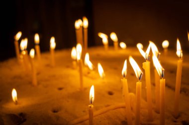 Bir grup yanan mum kumdan bir yatakta duruyor, titreyen alevleri sıcak, altın bir parıltı saçıyor. Balmumu, ateşin ışığı yansımayı ve bağlılığı sembolize eden barışçıl ve ruhani bir atmosfer yaratırken damlıyor.