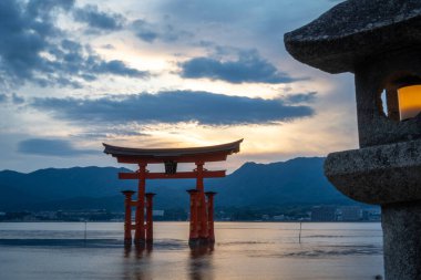 Itsukushima Tapınağı 'nın ünlü yüzen torii kapısı gün batımında sakin sularda duruyor. Taş fenerler ve uzak dağlarla çevrili.