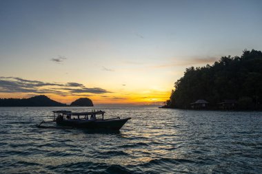 Gün batımında Bolilanga Adası, Togian takımadaları, Sulawesi, Endonezya sahillerinde sakin suları ve altın gökyüzü ile küçük bir tekne huzur içinde yüzer.