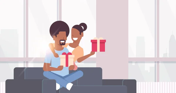Pár gazdaság jelenlegi keresztkötés ajándék dobozok afrikai amerikai férfi nő ül a kanapén ünnepli Nyaralás koncepció boldog lovers modern nappali belső vízszintes átfogó — Stock Vector