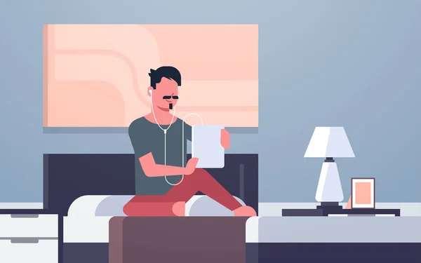Hombre escuchar audio libro a través de auriculares chico usando tableta sentado en la cama moderno dormitorio interior masculino carácter de longitud completa plana horizontal — Vector de stock