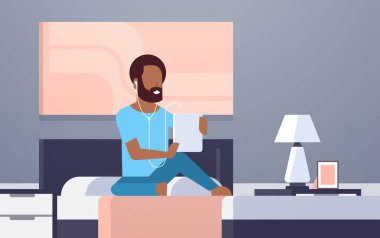 kulaklık Afrika americn adam yatak modern ev yatak odası iç erkek karakter tam uzunlukta üzerinde düz yatay oturan tablet kullanma yoluyla dinleme ses kaseti dostum