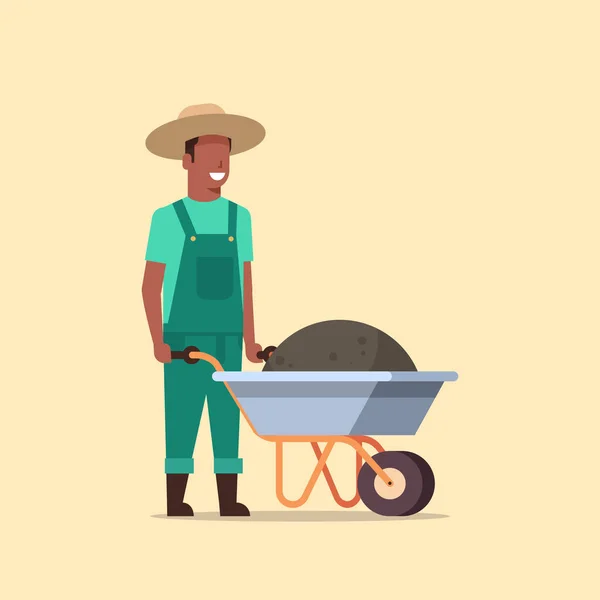 Tuinman mens duwen kruiwagen vol aarde compost African American mannelijke boer werken in de tuin dragen van overalls tuinieren concept volledige lengte — Stockvector