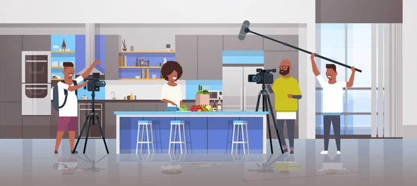 Χειριστές που χρησιμοποιούν βιντεοκάμερα εγγραφή τροφίμων blogger αφρικανική γυναίκα προετοιμασία νόστιμα πιάτα βιντεογράφους χρησιμοποιώντας επαγγελματικό εξοπλισμό μαγείρεμα ιστολόγιο έννοια κουζίνα εσωτερικό οριζόντια — Διανυσματικό Αρχείο
