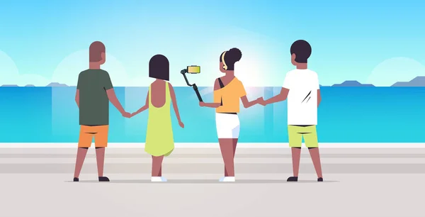 Menschen Gruppe am Strand mit Selfie-Stick Foto Smartphone-Kamera Sommerurlaub Konzept Rückansicht afrikanisch-amerikanische Freunde zusammen stehend Meer Hintergrund volle Länge horizontal — Stockvektor