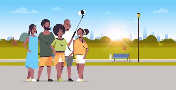 Gruppo di persone utilizzando bastone selfie scattare foto sulla fotocamera smartphone amici afro-americani in piedi insieme città parco urbano alba paesaggio sfondo full length orizzontale — Vettoriale Stock