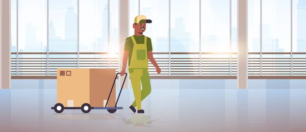 Mensajero en uniforme empujando carro con caja de cartón express delivery service concepto africano americano trabajador con mano camión moderno oficina hall interior plana longitud completa horizontal — Vector de stock