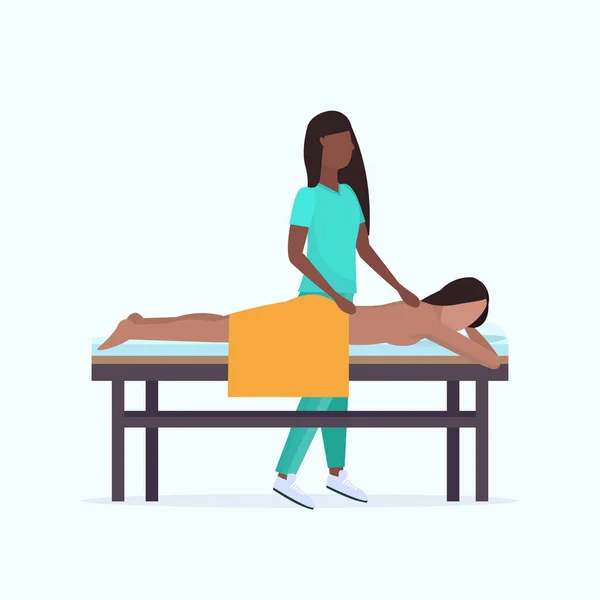 Joven chica teniendo espalda masaje africano americano masajista terapeuta en uniforme masaje paciente cuerpo mujer relajante acostado en cama spa salón tratamientos concepto de longitud completa — Vector de stock