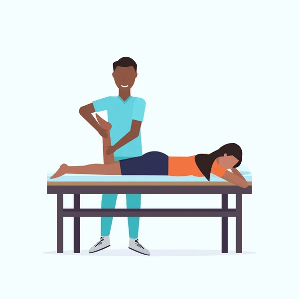 Mulher deitada na mesa de massagem Africano massagista americano massagista fazendo tratamento de cura massageando paciente ferido perna manual esporte fisioterapia conceito de reabilitação comprimento total —  Vetores de Stock