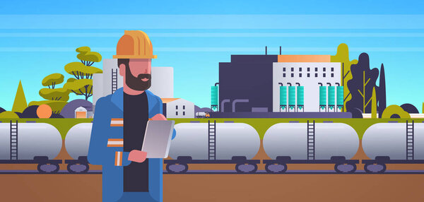 male engineer in helmet using tablet controlling train tanks with oil and fuel factory building industrial zone plant power station production technology concept horizontal
