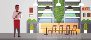 profesyonel garson elinde şarap şişesi tutan afro-amerikan erkek restoran işçisi kırmızı önlük alkol içki modern kafe iç düz tam uzunlukta yatay sunan