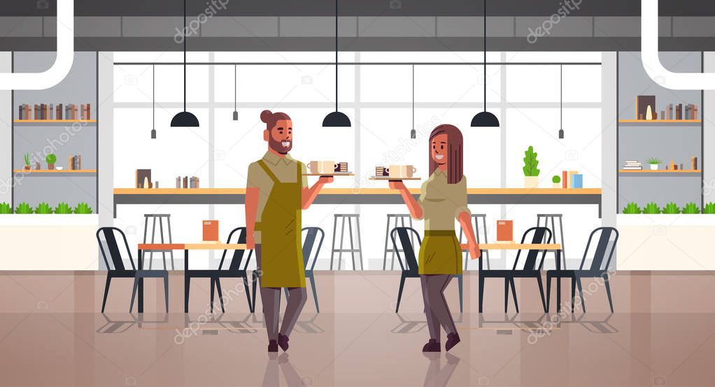 waiters couple carrying coffee and cakes on tray man woman cafe workers in aprons holding cups of cappuccino with desserts modern cafeteria interior full length horizontal