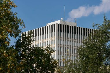 Çatıda dikey siyah çizgileri olan, yeşil ağaçlarla çerçevelenmiş, modern bir ofis kulesi, Saskatoon, Kanada.