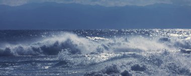 Rıhtımda denize karşı duran bir adam büyük bir dalgayla fırtına havasında su sıçratarak dövüyor.