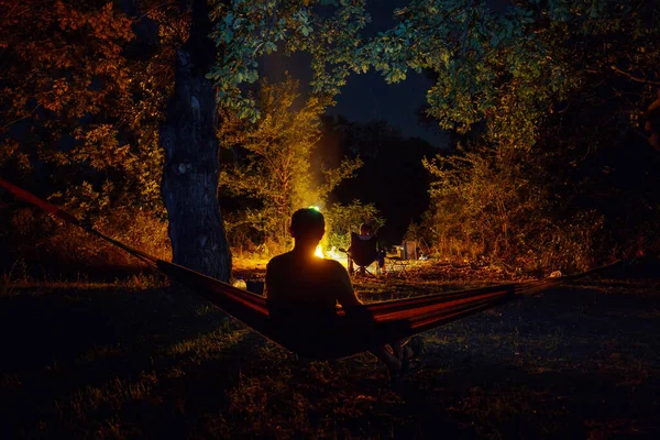 夜のキャンプで素晴らしいシーン 男でハンモックでたき火の背景 — ストック写真
