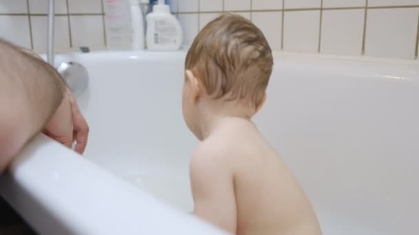 Adorable brown-eyed infant baby boy washing in bathtub with help of his parents — Stock Video