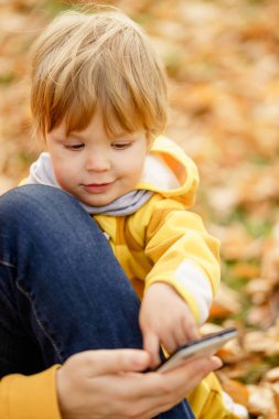 Mutlu çocuk gülüyor ve parkta sonbaharda annenin Smartphone ile oynuyor