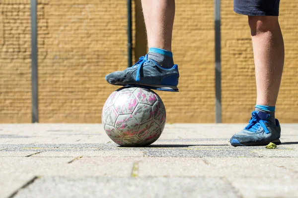 右の足の下でボロボロのボールとのストリート サッカーのピッチに立っている古いスニーカーを着て少年 — ストック写真