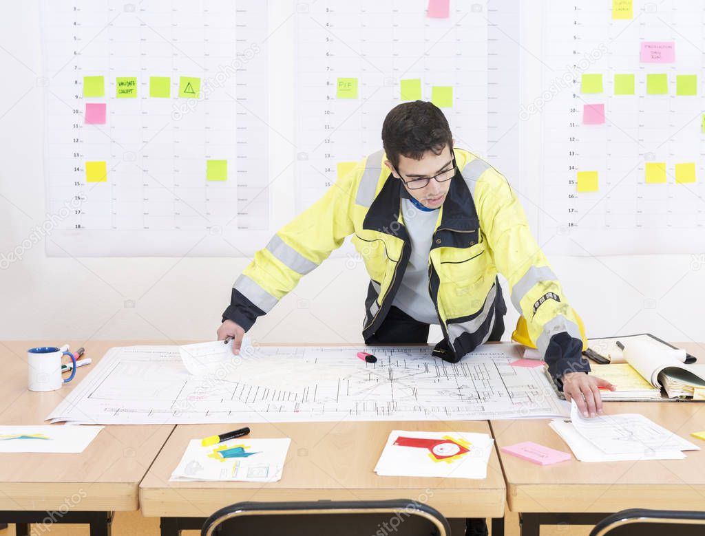 Construction worker checking design drawings in an office, wearing a saftety jacket