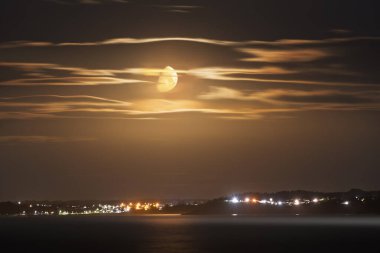 Kıyı İrlanda gece manzarası, parlak ay, çizgili bulutlar ve körfezin karşısındaki sıcak kasaba ışıkları