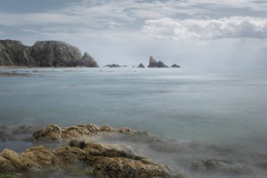 Gökyüzü sahnesi, altın-kahverengi kayalar, düzleştirilmiş Atlantik dalgaları, ve bulutlu bir günde engebeli burunlar.
