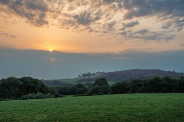Yeşil çayır, uzak tepe, sisli vadi, çiftlik evi ve parıldayan günbatımı olan İrlanda kırsalının göz hizasında manzarası.