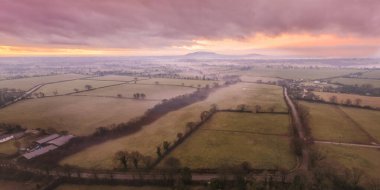 Şafak vakti İrlanda kırsalının atmosferik manzarası dalgalı tarlalar ve renkli gökyüzünün altındaki vadi sisi..