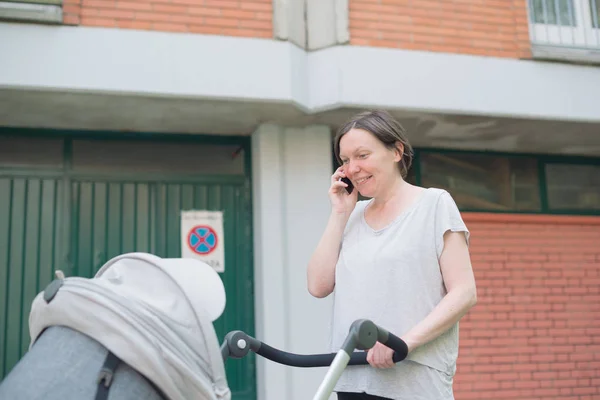 携帯電話で話していると カジュアルなスポーツ衣料の路上を歩きながらベビーカーで赤ちゃんを押して幸せな母 — ストック写真