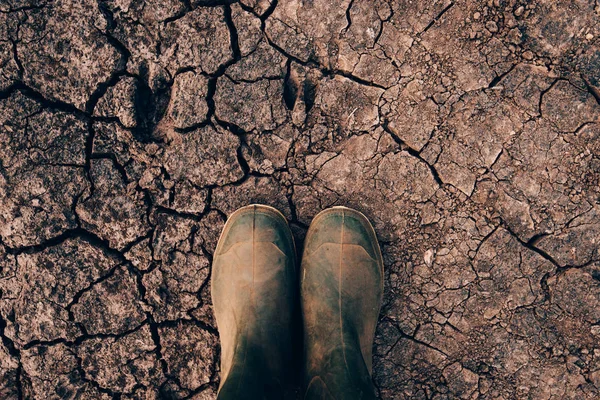 Farmer in rubber boots standing on dry soil ground, global warming and climate change is impacting crops growing and yield