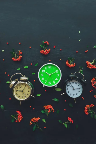 Ten past ten on three different alarm clock, flat lay top view minimal composition of timepieces placed on dark wooden background with decorative wild berry fruit arrangement