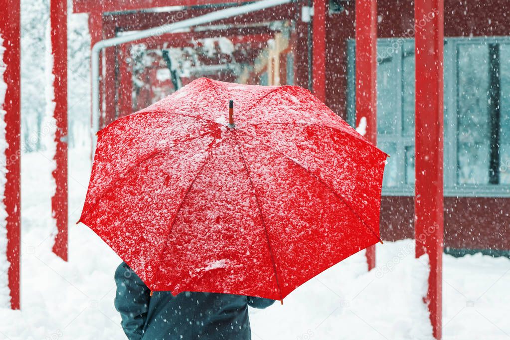Sad and alone woman under red umbrella walking in winter snow through urban environment
