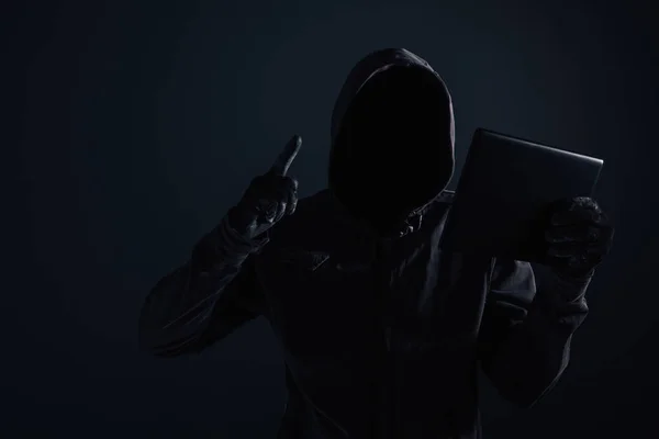 Hooded computer hacker with obscured face using digital tablet in cybercrime and cybersecurity concept, low key with selective focus