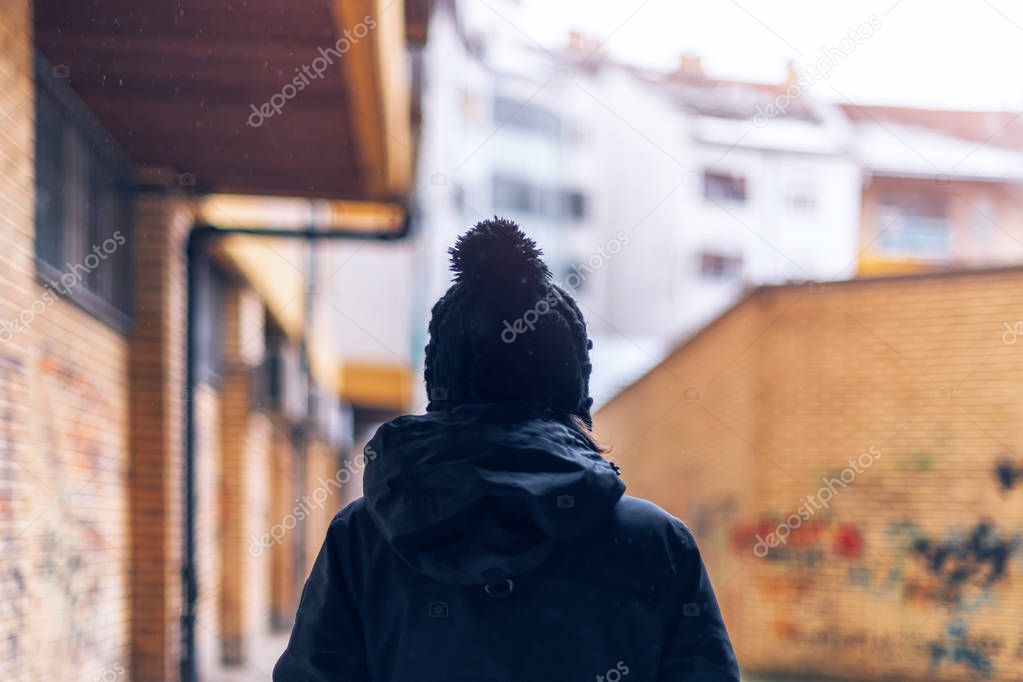 Rear view of casual adult woman walking on urban city street during the light snowfall in winter