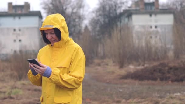 手持ちのスローモーション映像 雨の日の屋外の携帯電話上に黄色のレインコート テキスト メッセージで女性 — ストック動画