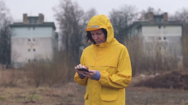 手持ちのスローモーション映像 雨の日の屋外の携帯電話上に黄色のレインコート テキスト メッセージで女性 — ストック動画